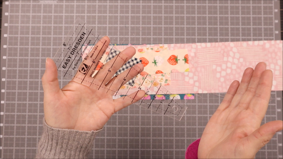 Two hands hold a clear plastic Dresden Plate ruler labeled "Easy Dresden" above a pink, patterned fabric strip on a gray cutting mat with a grid, ready to create beautiful quilt pieces.