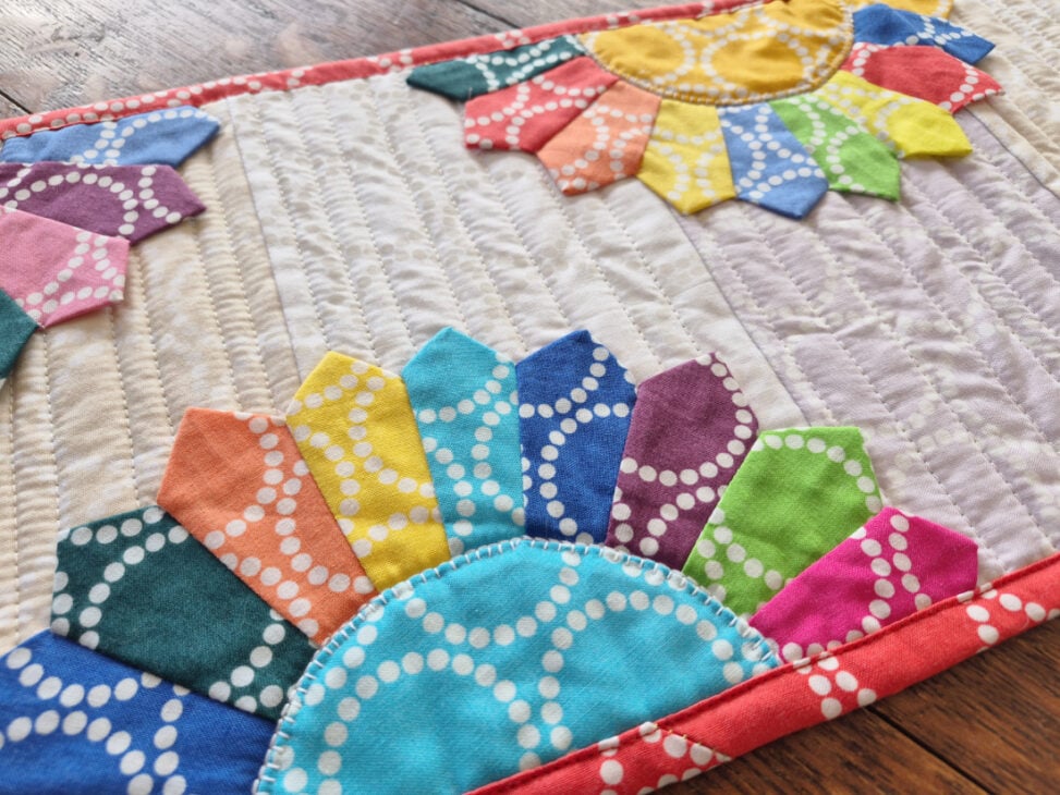 Dresden Plates Table Runner & Mug Rug - a Pattern Set 4 A quilted fabric piece features colorful Dresden Plates patchwork designs with dotted patterns on a neutral background, bordered by red fabric. The vibrant patches resemble flower petals. The quilt rests on a wooden surface.