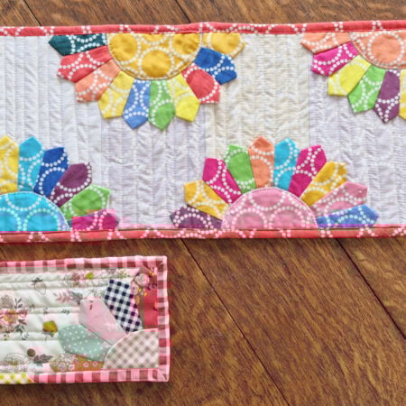 A colorful quilted table runner with vibrant Dresden Plates and a small quilted piece featuring a house and floral design lie on a wooden floor—perfect inspiration for your next Pattern Set or How To project.