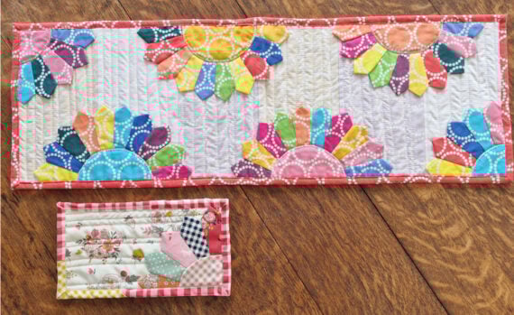 A colorful quilted table runner with vibrant Dresden Plates and a small quilted piece featuring a house and floral design lie on a wooden floor&mdash;perfect inspiration for your next Pattern Set or How To project.