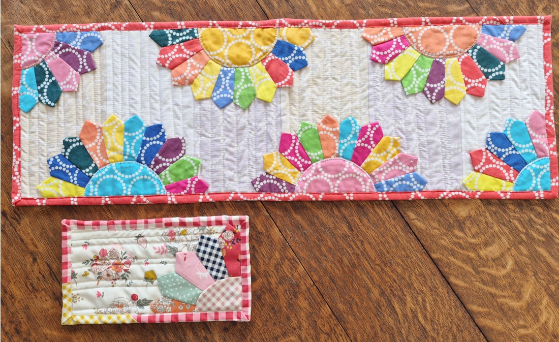 A colorful quilted table runner with vibrant Dresden Plates and a small quilted piece featuring a house and floral design lie on a wooden floor&mdash;perfect inspiration for your next Pattern Set or How To project.