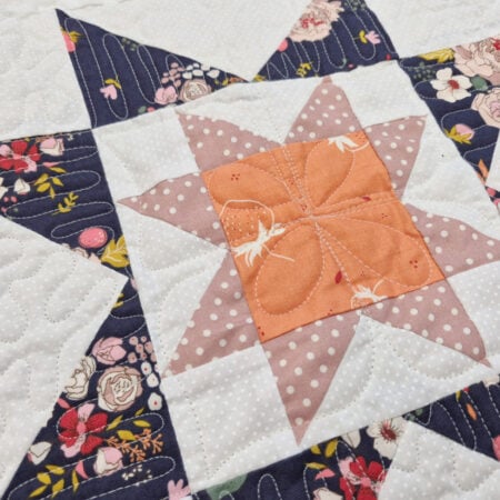 A close-up of a quilt featuring a star pattern with orange, pink, and white polka dot fabrics in the center, surrounded by navy floral and white background fabrics. Stitching details are visible throughout the quilt.