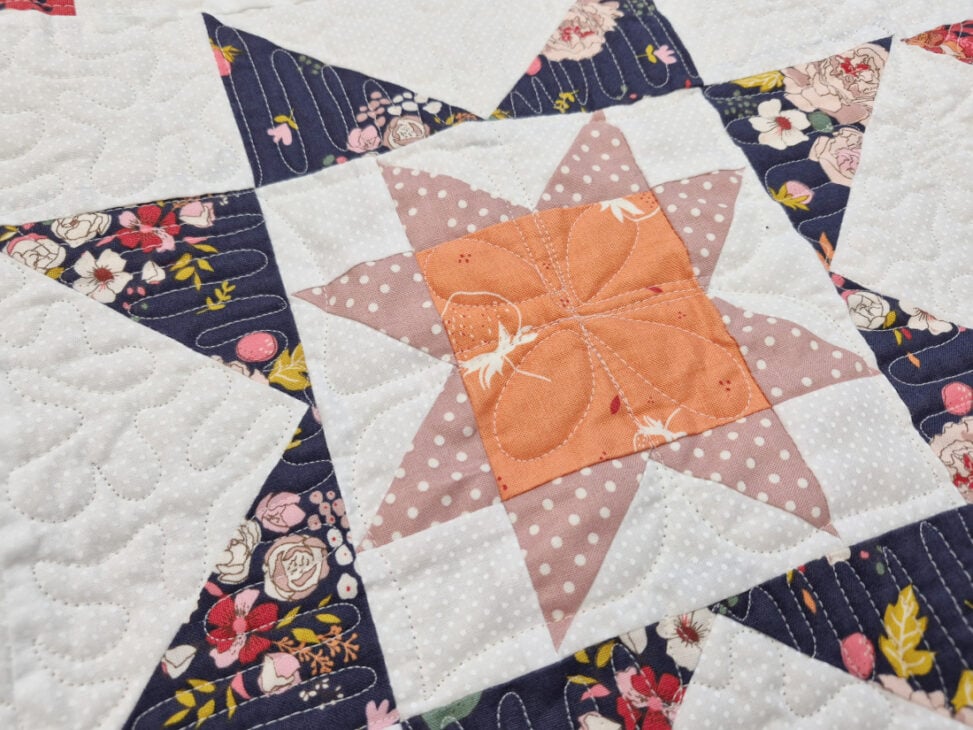 A close-up of a quilt featuring a star pattern with orange, pink, and white polka dot fabrics in the center, surrounded by navy floral and white background fabrics. Stitching details are visible throughout the quilt.