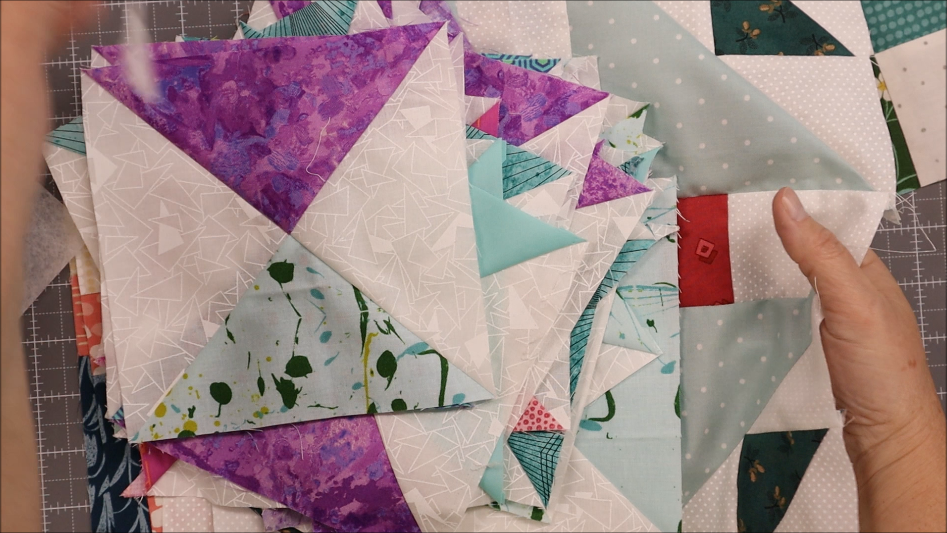 A person holds several quilt blocks made from colorful fabric triangles and patterns in shades of purple, teal, white, and printed designs&mdash;perfect for crafting a sew pen holder or a unique pen bookmark holder&mdash;arranged on a cutting mat.