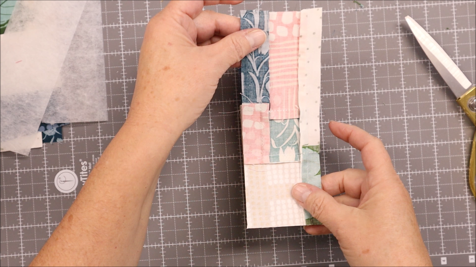 A person arranges strips of patterned fabric and elastic on a gray cutting mat, preparing to sew or quilt. Scissors and extra fabric pieces lie nearby, ready for the next creative step.