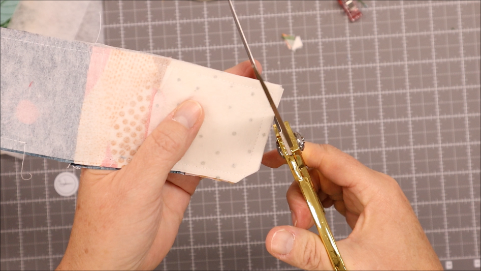 A person uses gold scissors to trim the corner of fabric pieces sewn together with elastic, on a cutting mat with a grid pattern&mdash;an essential step in learning how to sew projects like a sew pen bookmark holder.