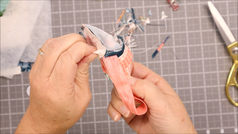 A person turns a small fabric pouch right side out on a gray grid cutting mat, preparing to sew a Pen Bookmark Holder with elastic, surrounded by fabric scraps and gold-handled scissors.