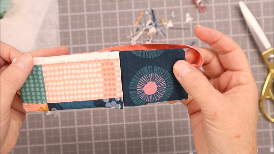 Two hands hold a fabric strip, sewn with different patterns like pink polka dots and blue circles, perfect for creating an elastic Pen Bookmark Holder. Scissors and fabric scraps rest on a gridded cutting mat in the background.