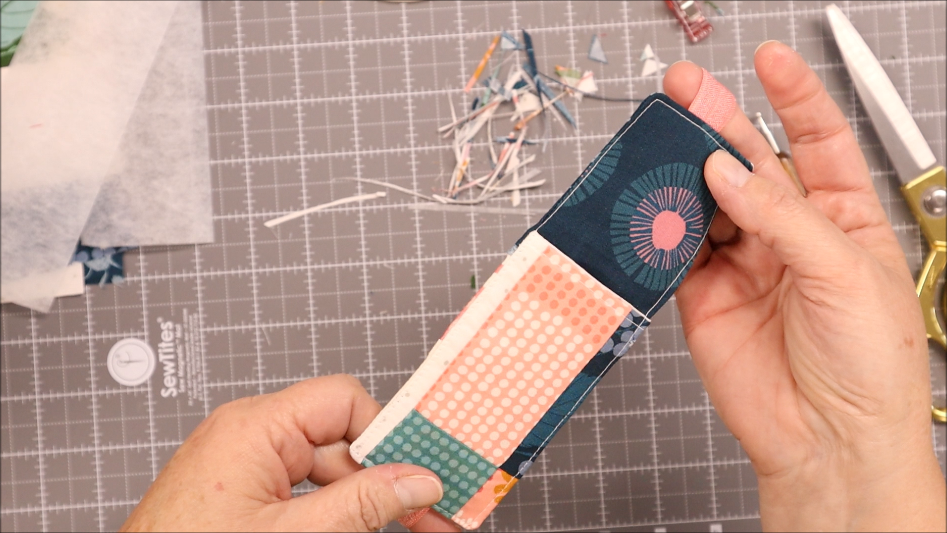 A person holds a sewn fabric pen bookmark holder with colorful patterns over a gridded cutting mat. Scissors, fabric scraps, and crafting materials are visible in the background.