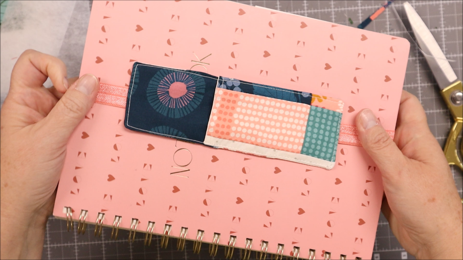 A person holds a pink spiral notebook with cut-out hearts and the word "LOVE." A colorful elastic bookmark is wrapped around the cover, and scissors rest nearby on a crafting mat.