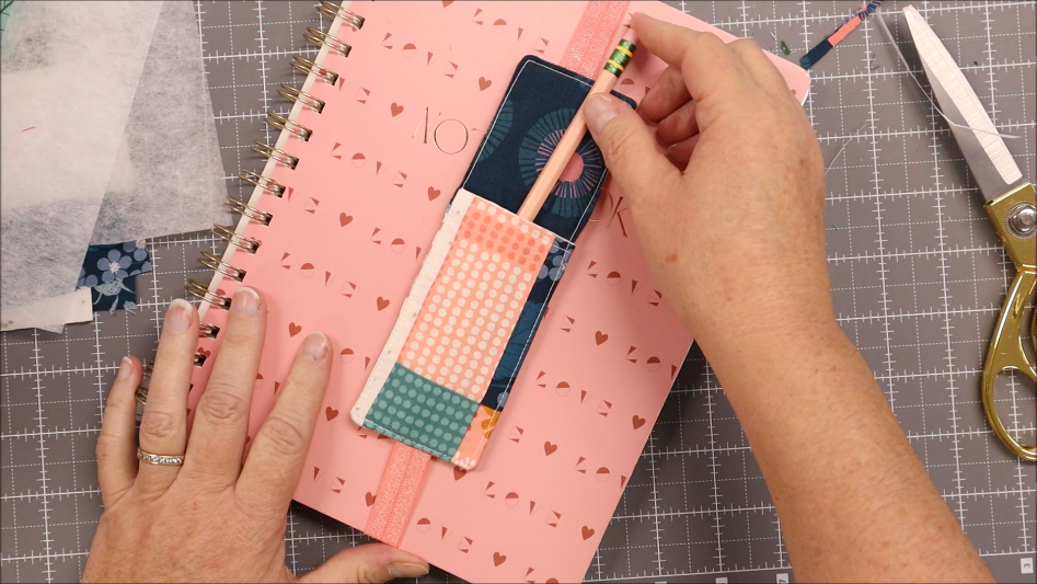 A person places a pencil into a colorful fabric pen bookmark holder attached to a pink notebook with small heart cutouts. Scissors and crafting materials are nearby on a gray grid work surface.