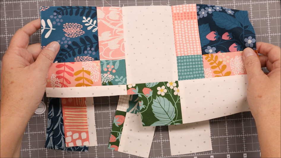 Two hands hold a partially assembled quilt block made of colorful patterned fabric squares and white strips, ready to sew. An elastic pen bookmark holder lies nearby on the cutting mat with measurement grid lines.