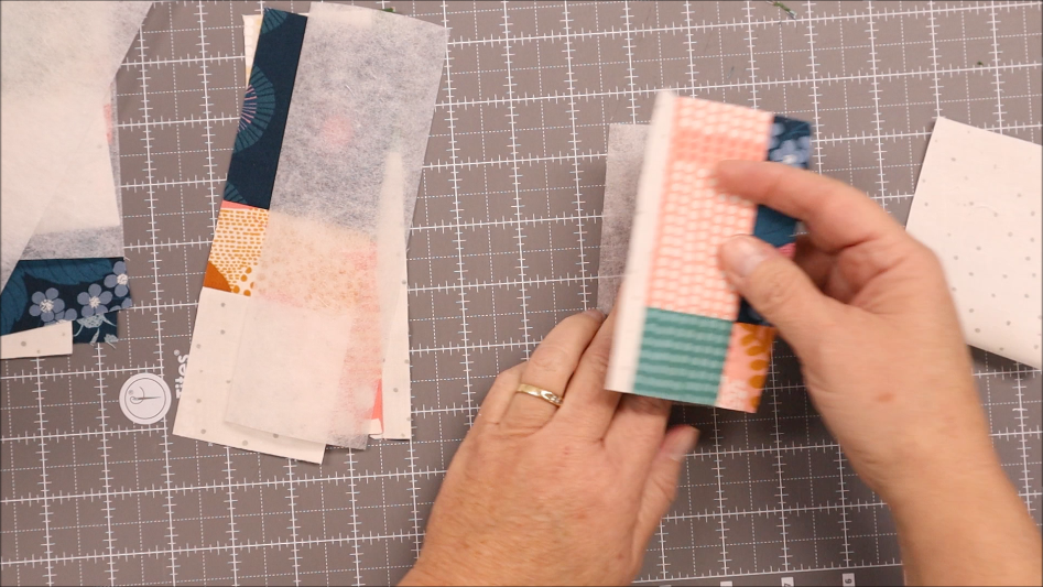 A person holds a stack of small, colorful fabric strips, preparing to sew them into elastic Pen Bookmark Holders on a gray gridded cutting mat, with more fabric strips and materials lying nearby.