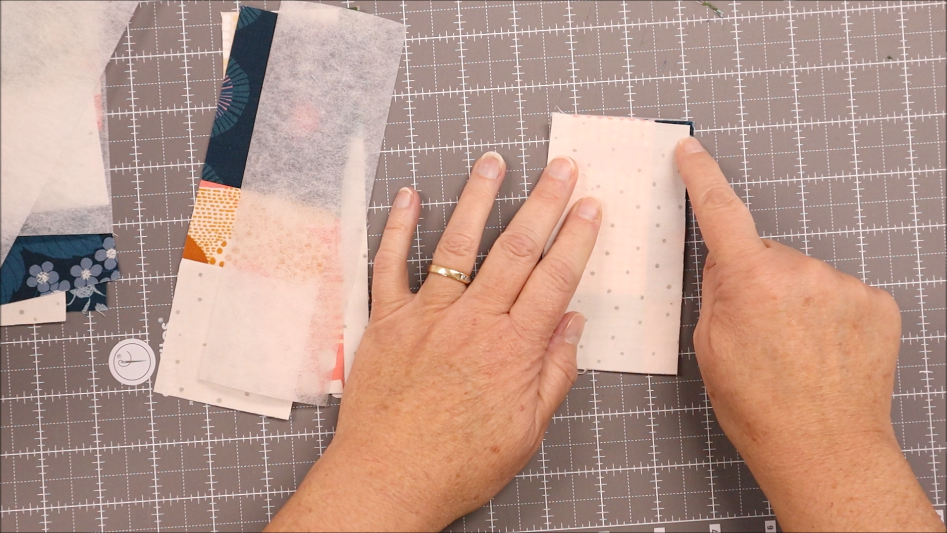 Two hands holding and pointing at a rectangular piece of white fabric with small dots, placed on a cutting mat next to other fabric pieces, strips of interfacing, and elastic for a Sew pen bookmark holder tutorial.