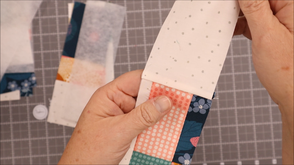 A person holds two quilt fabric strips, one white with gray dots and one multicolored with patterns, aligning the edges on a cutting mat background&mdash;perfect for your next sew project like an elastic pen bookmark holder.