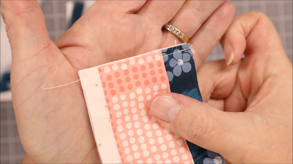 A hand holds a piece of quilted fabric with pink, white, and blue patterned strips, showing a close-up of the neat stitching and an elastic edge&mdash;perfect to sew into a Pen Bookmark Holder. The person is wearing a gold ring.