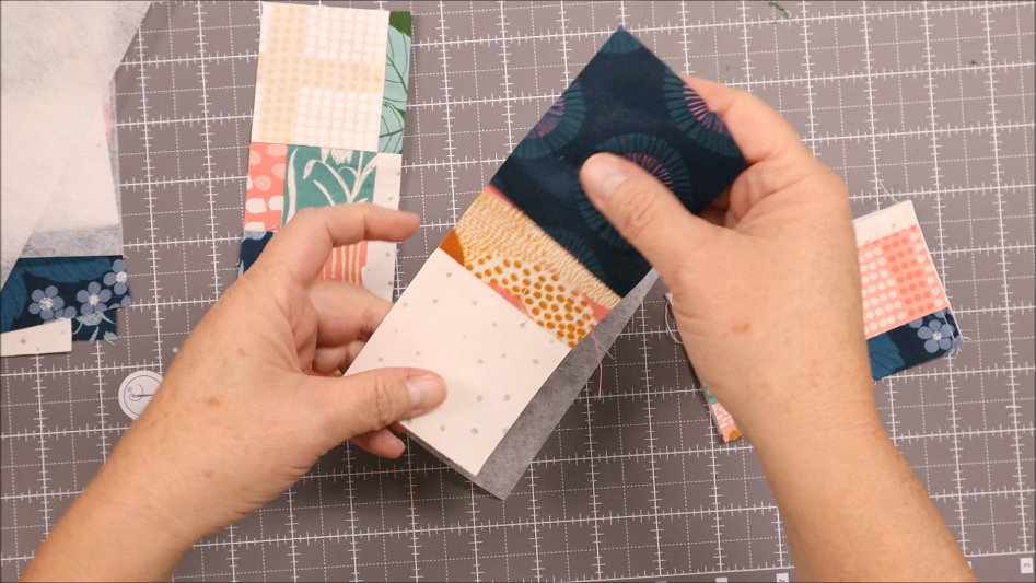 A person holds a colorful patchwork piece over a gray cutting mat with gridlines, surrounded by fabric pieces and cut squares&mdash;perfect materials to sew a Pen Bookmark Holder or craft your own DIY Pen Bookmark.