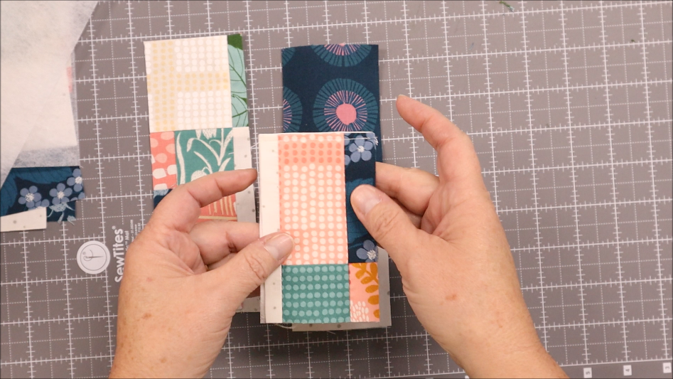 A person holds pieces of colorful patterned fabric, preparing to sew them together into a sew pen holder, on a gray cutting mat marked with a grid.