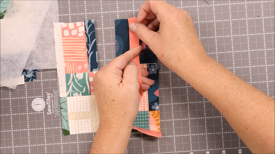 Hands are aligning and pressing a strip of colorful patterned fabric on a patchwork piece atop a gray cutting mat with grid lines in this Quick Sewing Tutorial, perfect for creating an Elastic Pen Holder.