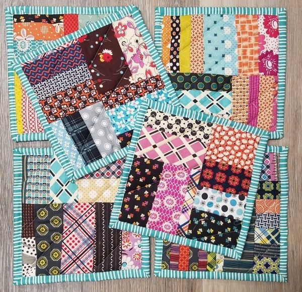 Four colorful, patchwork quilted coasters, perfect as mini hot pads, feature various patterned fabric strips and are bordered by turquoise and white striped edges, all arranged overlapping on a wooden surface.