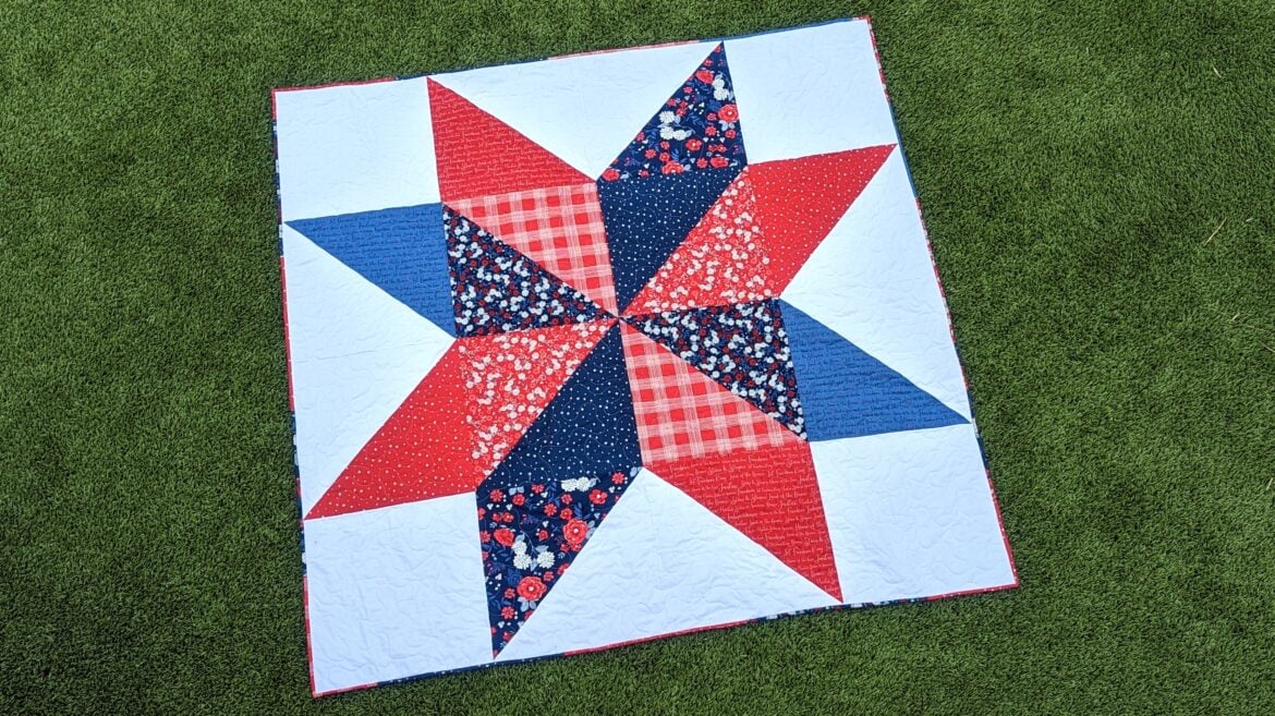 10 Patriotic Flag Projects to Sew Before the 4th of July 16 A colorful quilt with a large star pattern in red, navy blue, white, plaid, and floral prints lies flat on green grass. Perfect for 4th of July celebrations or Patriotic Flag Projects, this square quilt features various fabric textures and designs.