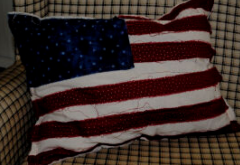 10 Patriotic Flag Projects to Sew Before the 4th of July 5 A rectangular pillow with an American flag design, perfect for 4th of July decor, rests on a checkered armchair. The flag pattern features dark blue with white dots and red and white stripes—ideal inspiration for patriotic flag projects.