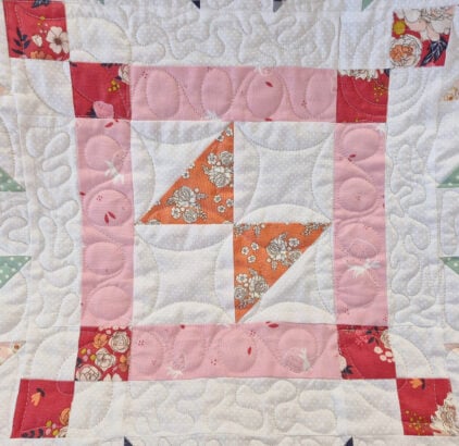 A quilt block for the March 2026 BOM featuring a central pinwheel in orange floral fabric, surrounded by white fabric, a pink border, and red floral corner squares, all highlighted with detailed quilted swirl stitching.