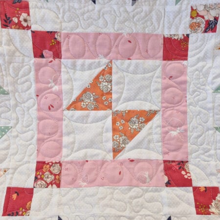 A quilt block for the March 2026 BOM featuring a central pinwheel in orange floral fabric, surrounded by white fabric, a pink border, and red floral corner squares, all highlighted with detailed quilted swirl stitching.