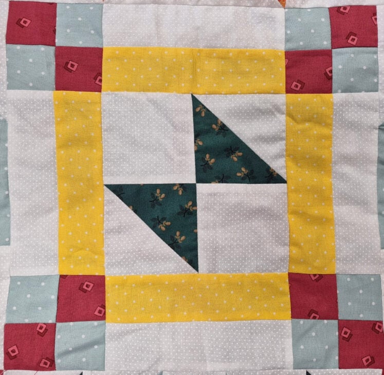 A quilt block for the March 2026 BOM featuring a geometric pattern with yellow, white, blue, and red fabrics, plus a dark green floral print in the center, arranged in triangles and squares with a patchwork border.