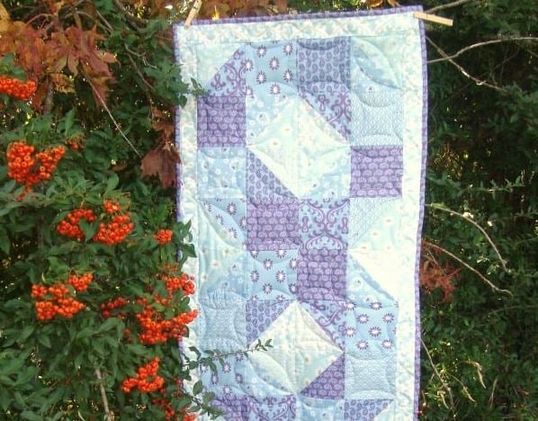 Easy Spring Table Runner Patterns You Can Sew in a Day 2 A patchwork quilt in shades of purple, blue, and white hangs outdoors on a leafy bush with clusters of bright red-orange berries—an inspiring sight for easy sewing or spring table runner patterns you can sew in a day.