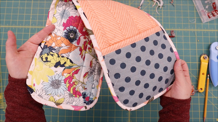 A person holds two colorful quilted hot pad fabric pieces with rounded edges; one side has a bright floral pattern, and the other features orange chevrons and blue polka dots. A green cutting mat and sewing tools are in the background.