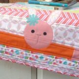 A colorful quilted sewing machine cover, decorated with a cute pink bunny face and vibrant stripes, adds charm to any space. Perfect for easy sewing, it brightens your room while books stack neatly on top of the covered machine.