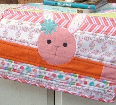 A colorful quilted sewing machine cover, decorated with a cute pink bunny face and vibrant stripes, adds charm to any space. Perfect for easy sewing, it brightens your room while books stack neatly on top of the covered machine.