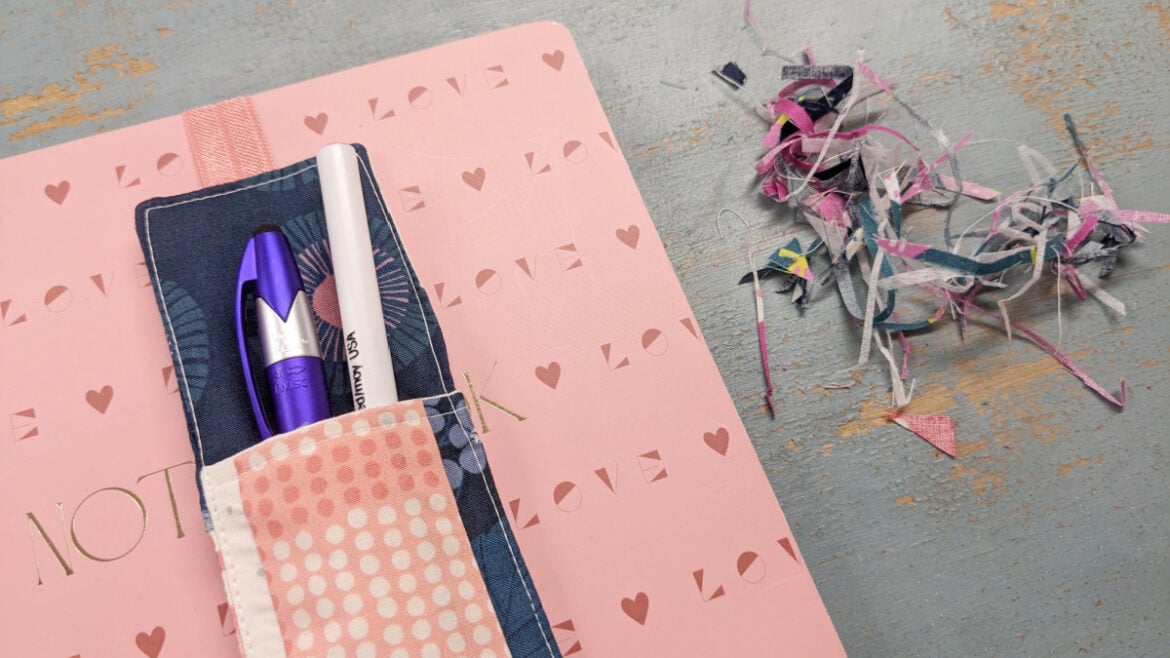 A pink notebook with "LOVE" and heart patterns, a sewn fabric pen bookmark holder with elastic, two pens, and a pile of colorful paper scraps on a light blue wooden surface.
