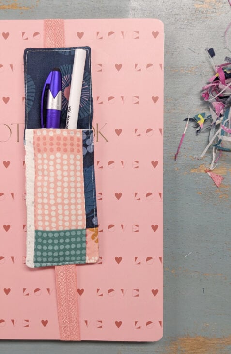 A pink notebook with "LOVE" and heart patterns features a sewn elastic pen bookmark holder attached to the cover, holding a purple pen and white marker. Colorful fabric scraps are on the right on a wooden surface.