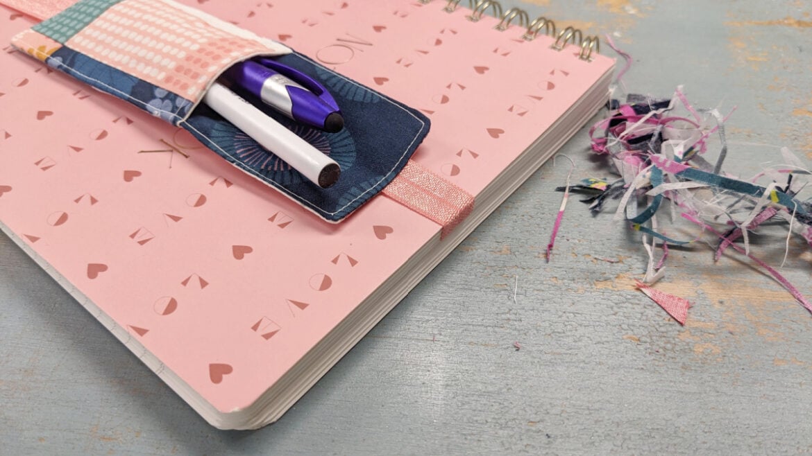 A pink spiral notebook with &ldquo;LOVE&rdquo; and heart shapes on the cover sits on a gray surface. Two pens are tucked in a DIY bookmark pen holder on the notebook, with scraps of colorful paper and fabric nearby.
