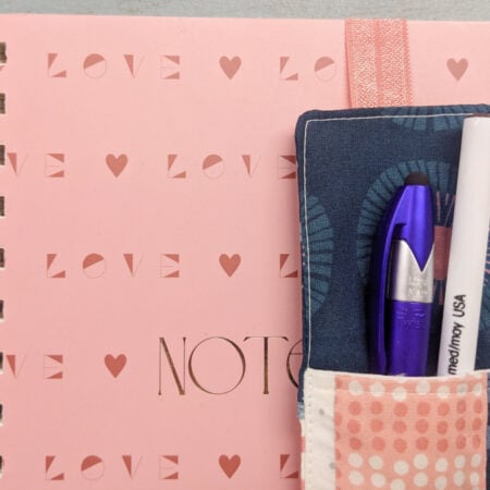 A pink spiral notebook with "LOVE" in gold, featuring a sewn fabric pen bookmark holder with elastic, containing a blue and a white pen. The notebook rests on a light-colored surface.