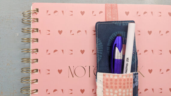 A pink spiral notebook with "LOVE" in gold, featuring a sewn fabric pen bookmark holder with elastic, containing a blue and a white pen. The notebook rests on a light-colored surface.