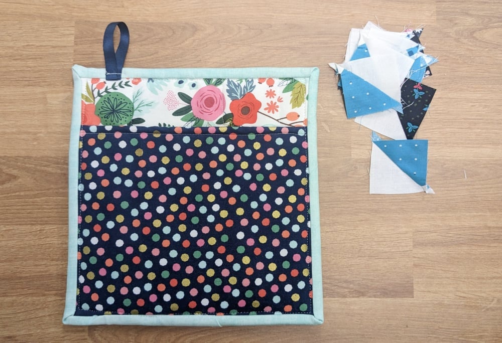 A square quilted hot pad with a floral and polka dot design lies on a wood surface next to a small pile of blue, black, and white fabric triangles.