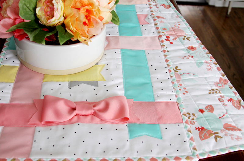 Easy Spring Table Runner Patterns You Can Sew in a Day 7 An easy sewing project, this quilted table runner features pastel ribbons and bow designs—perfect for spring table runner patterns. It sits on a wooden table with a white vase of colorful artificial flowers on top.