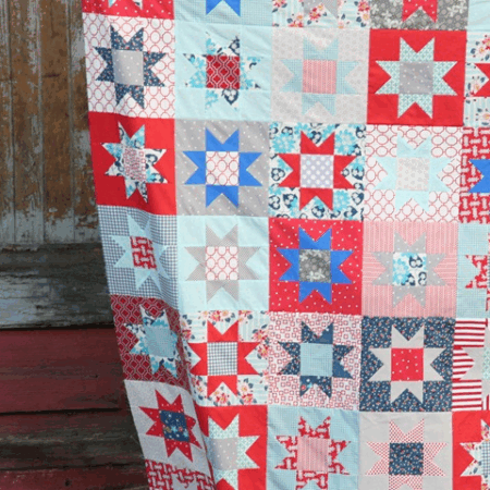 A colorful patchwork quilt featuring red, blue, and white star patterns, perfect for patriotic flag projects or to sew for the 4th of July, hangs in front of a rustic wooden wall with peeling paint.