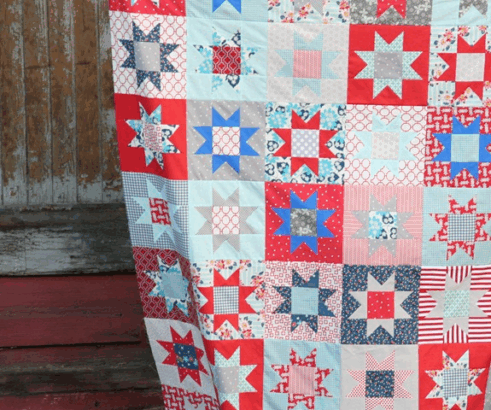 A colorful patchwork quilt featuring red, blue, and white star patterns, perfect for patriotic flag projects or to sew for the 4th of July, hangs in front of a rustic wooden wall with peeling paint.