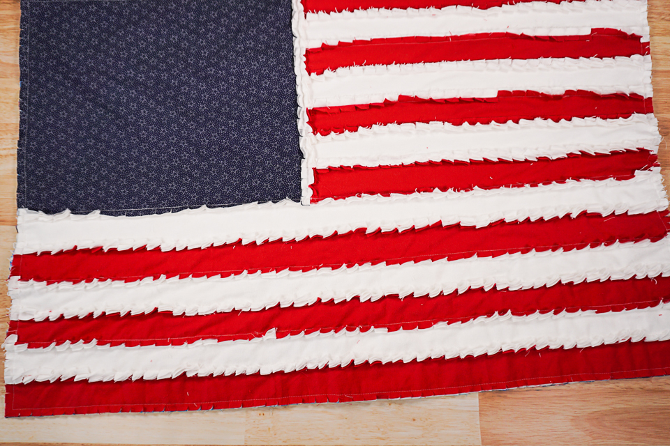10 Patriotic Flag Projects to Sew Before the 4th of July 14 A handmade American flag made from fabric strips, perfect for 4th of July or other patriotic flag projects, featuring a blue star section and sewn ruffled red and white stripes, all displayed on a wooden surface.