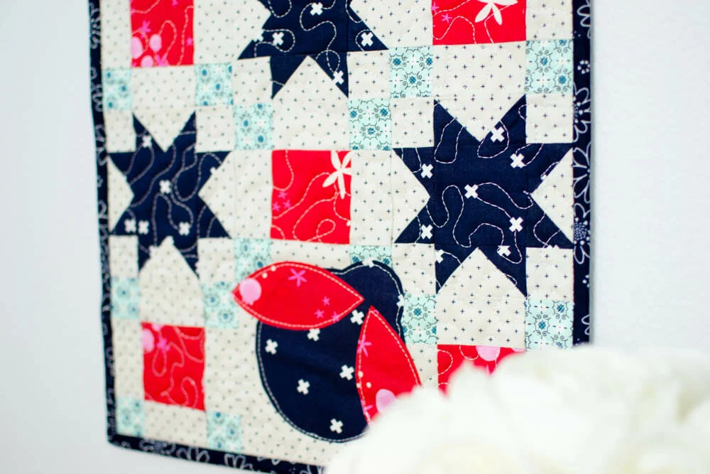 10 Patriotic Flag Projects to Sew Before the 4th of July 15 Colorful quilt with a pattern of navy stars, light blue squares, and a red and navy appliqué bird—perfect for 4th of July or patriotic flag projects—set against a white background with a blurred white flower in the foreground.