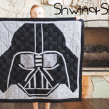 A child stands indoors holding a quilt with a large Darth Vader mask design—perfect for Star Wars fans. A stone fireplace is in the background. The words "Shwin & Shwin" appear in the top right corner.