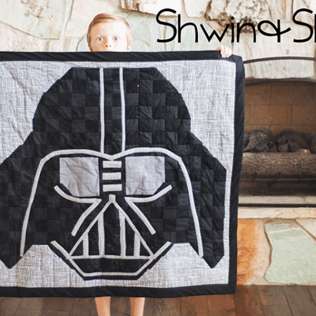 A child stands indoors holding a quilt with a large Darth Vader mask design—perfect for Star Wars fans. A stone fireplace is in the background. The words "Shwin & Shwin" appear in the top right corner.