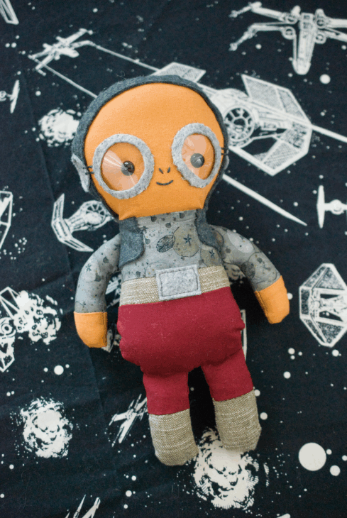 A handmade cloth doll with an orange face, button eyes, silver goggles, gray shirt with space patterns, red pants, and beige boots lies on a black Star Wars-themed fabric&mdash;perfect inspiration for Star Wars quilt projects loved by fans.
