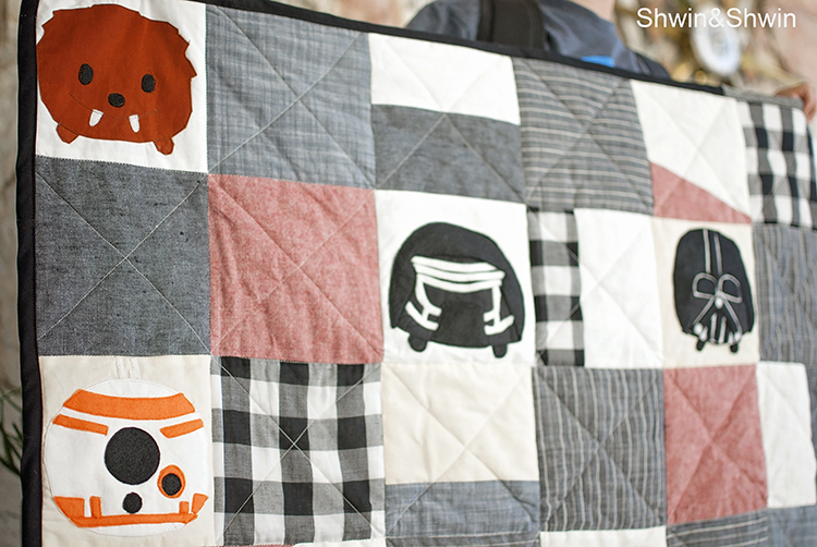 A quilt featuring a patchwork of gray, white, black, and rust-colored squares, with some displaying stylized Star Wars character faces&mdash;perfect inspiration for Star Wars quilt projects or as a cozy gift for Star Wars fans.