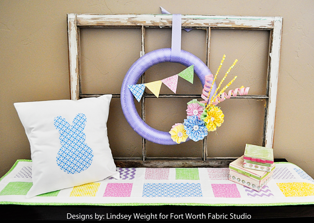 Easy Spring Table Runner Patterns You Can Sew in a Day 8 A decorative spring scene with a white pillow featuring a blue bunny silhouette, an easy sewing projects pastel patchwork table runner, a purple wreath with flowers and bunting, and stacked pastel boxes, all set against an old window frame.