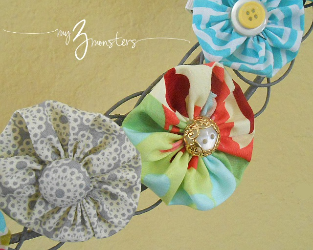 A close-up of three colorful fabric yo-yo flowers, each with a decorative button center, attached to a wire frame against a yellow background&mdash;an inspiring idea for fun fabric yo-yo projects. The words "my 3 monsters" are written in white script.